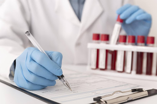 Frontal view of gloved hand holding pen over an official form attached to a clipboard and placed beside vials containing blood samples.