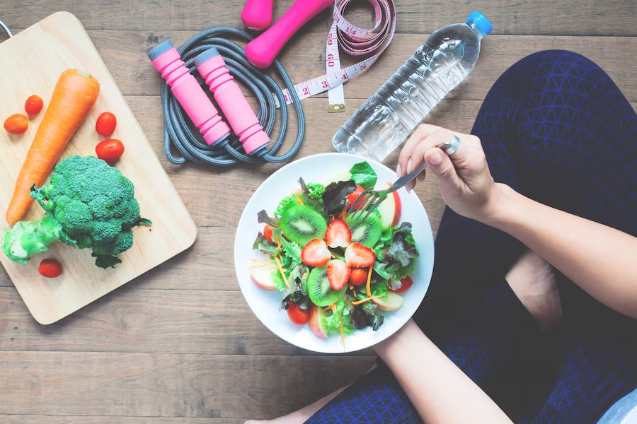 The top view of a person holding a plate of fruits while vegetables, skipping rope, a measuring tape and a bottle of water lay beside her.